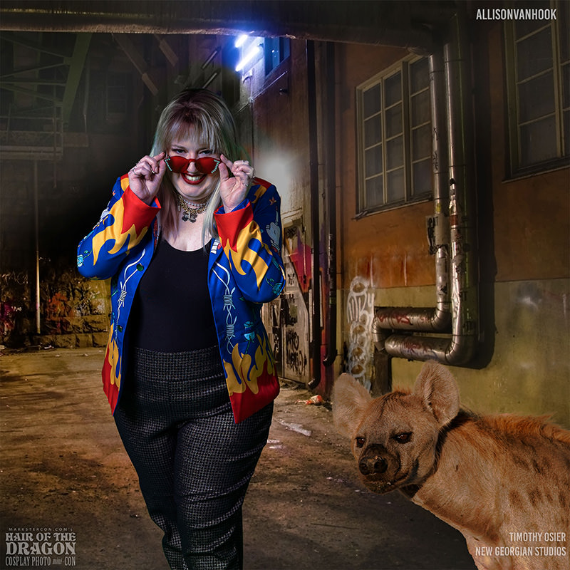 A woman wearing a colorful jacket with flame patterns, black top, and checkered pants is playfully pulling down red sunglasses while smiling. Behind her to the right, a hyena stands in a dimly lit, graffiti-covered urban alley.