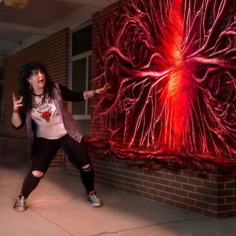 A person with curly dark hair, wearing a white Hellfire Club t-shirt, black ripped jeans, and colorful sneakers, stands with a surprised expression and raised hands in front of a brick wall. The wall features a glowing red portal with twisted roots extending outward, emitting bright red light.