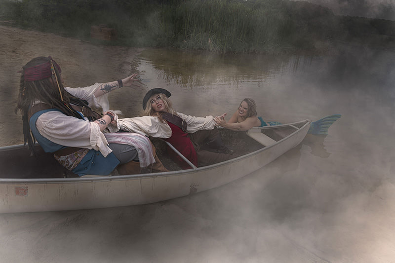 Two people dressed as pirates struggle in a canoe while a woman dressed as a mermaid pulls one pirate's hand from the water, surrounded by thick mist on a lake shore