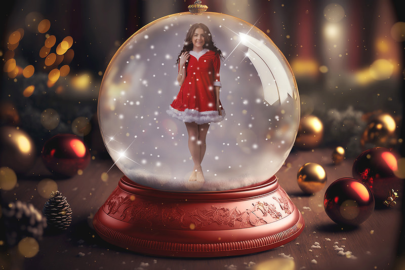 A woman wearing a red and white Santa dress stands barefoot inside a large, ornate snow globe with falling snowflakes, surrounded by Christmas ornaments and pine cones on a wooden surface with warm glowing lights in the background.