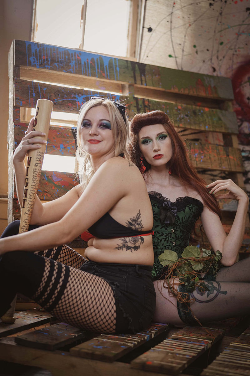Two women sitting back to back on a wooden pallet. The woman on the left has blonde hair, dramatic blue eye makeup, tattoos on her side and arm, black fishnet stockings, a black bra top, and holds a wooden bat. The woman on the right has long red hair, green eye makeup, a green corset with black lace, and holds a vine with green leaves. The background features a brightly lit window and a paint-splattered wooden pallet.