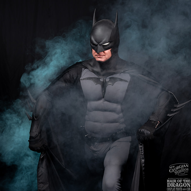 Person dressed as Batman in a detailed costume with cape spreading, surrounded by blue-gray smoke on a dark background