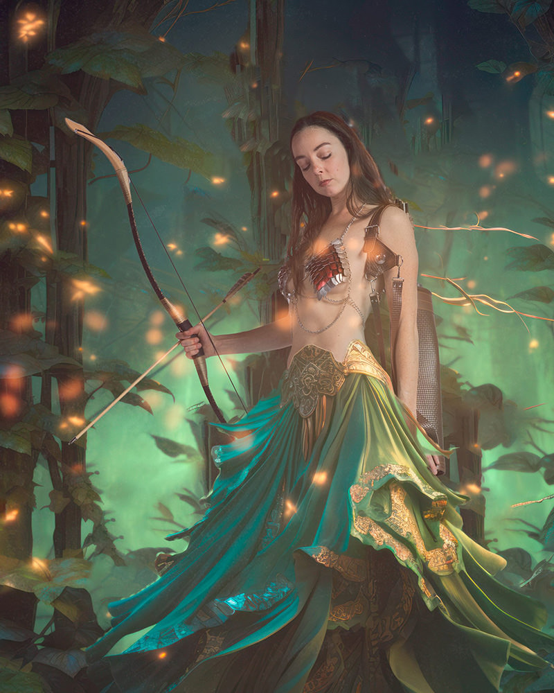 A woman dressed in fantasy armor with a metallic scale bra and flowing green skirt stands in a glowing forest holding a bow with a drawn arrow. Firefly-like lights float around her.