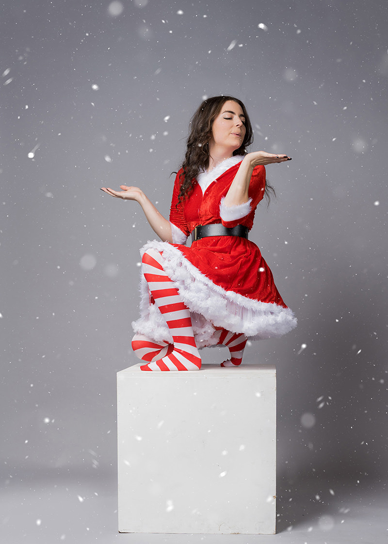 Young woman dressed in a red and white Santa-inspired outfit with a fur trim, red and white striped tights, kneeling on a white cube, with eyes closed and hands raised as if catching snowflakes, against a gray background with falling snow.