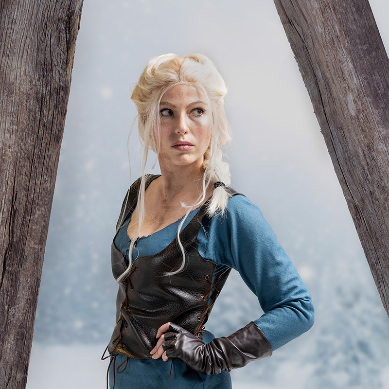 Young woman with long blonde braided hair wearing a blue medieval-style dress with a black leather vest and fingerless gloves, standing between two large wooden beams with a blurred light background.