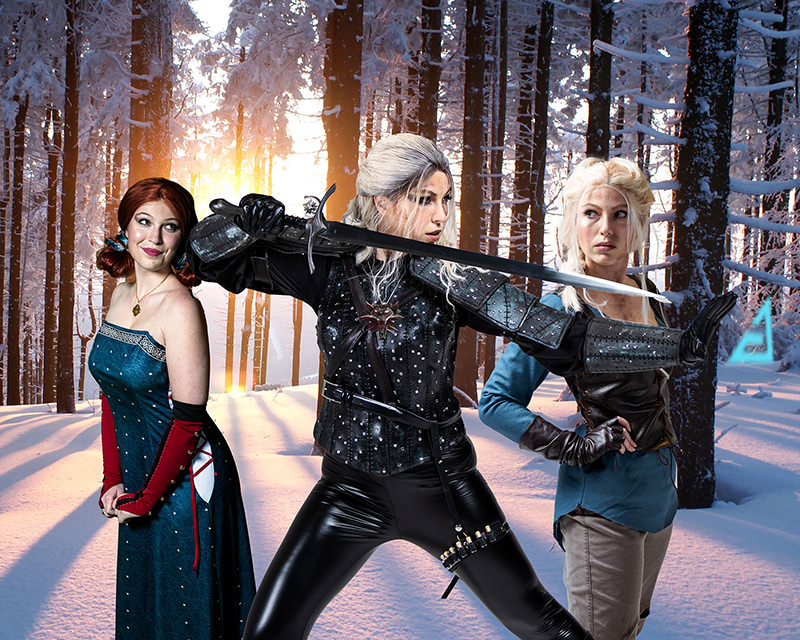 Three women dressed in fantasy warrior costumes stand in a snowy forest with tall trees and a setting sun in the background. The central figure with white hair holds a sword ready for battle, the woman on the left in a blue gown smiles, and the woman on the right in a medieval outfit poses confidently.