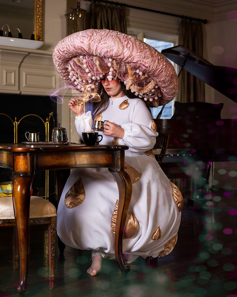 Person sitting barefoot at a wooden table wearing a large, ornate pink mushroom-shaped hat and a white dress with gold patches, holding a small cup and pouring liquid into a black cup, with a fireplace and piano in the background