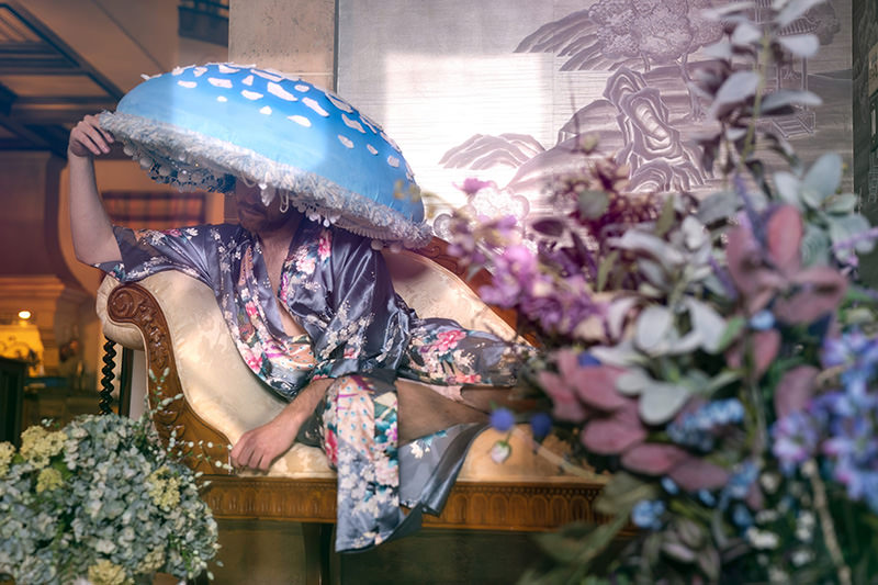 Person reclining on an ornate cream and wood chaise lounge wearing a silky floral robe and holding a large blue mushroom-shaped hat partially covering their face. There are floral arrangements in the foreground and a decorative wall behind them.