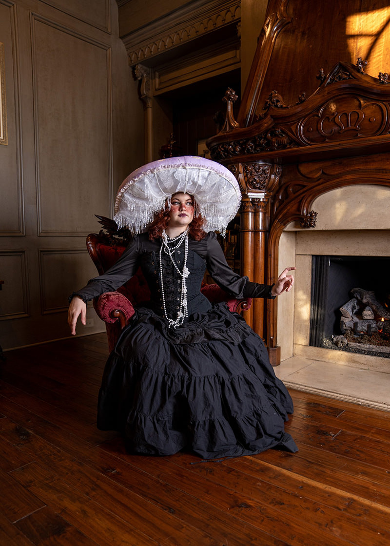 A person dressed in a Victorian-style black dress and a large white lace hat, sitting on a red upholstered chair next to an ornate wooden fireplace, with wooden floor and panelled walls in the background.
