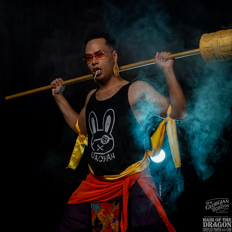 Person wearing red sunglasses, black tank top with bunny logo, yellow and orange fabric accessories, holding a large golden staff across shoulders with blue smoke swirling around against a dark background