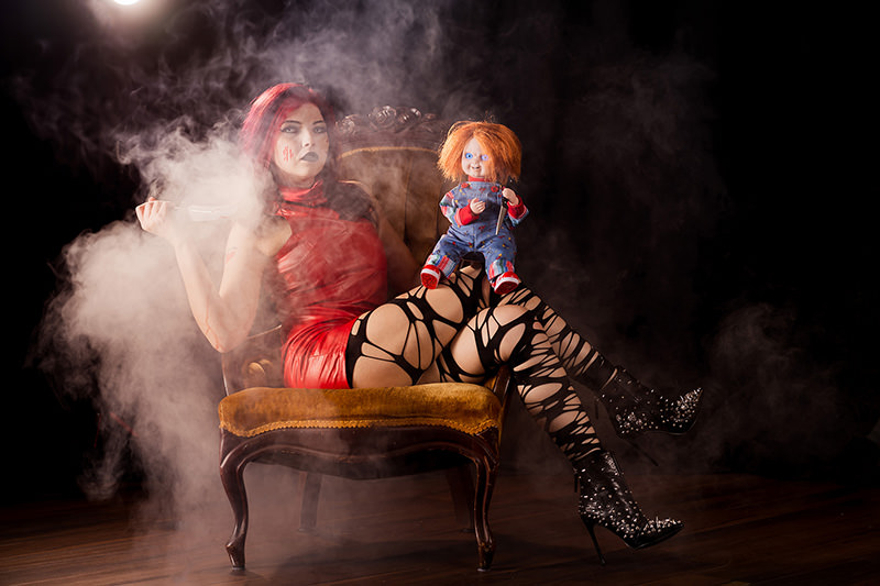 Woman with long red hair, wearing a red dress and black patterned stockings, sitting sideways on a vintage chair with smoke around her. She holds a knife in one hand while a doll with orange hair, blue eyes, and a striped outfit sits on her lap. The background is dark and smoky.