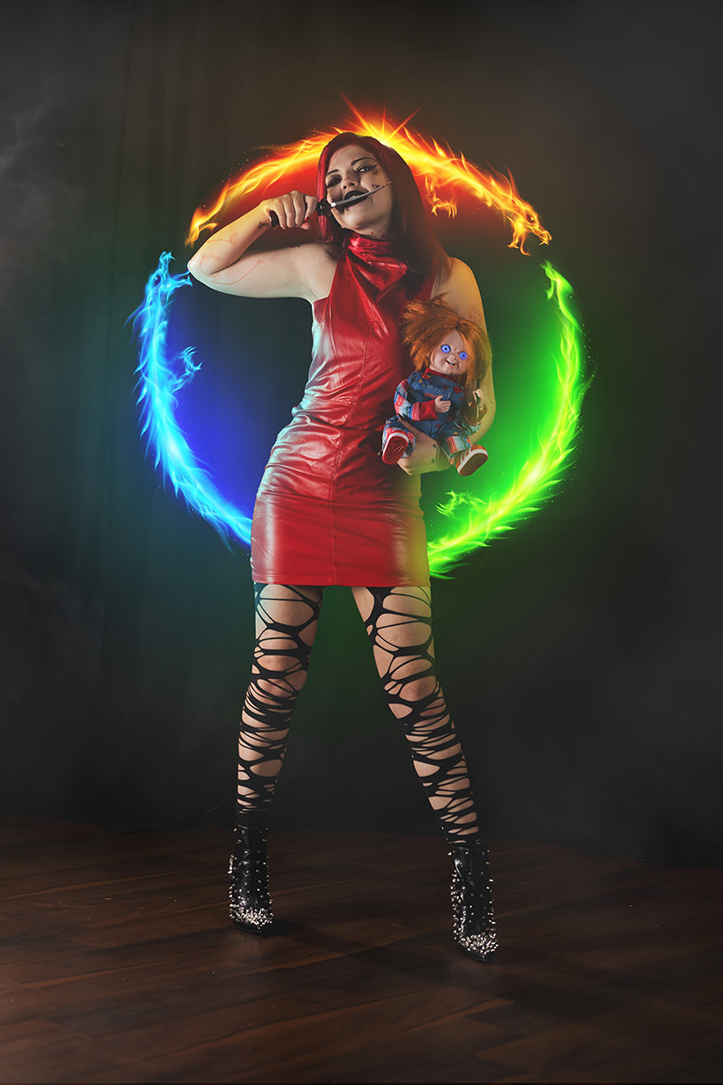 Woman in a red leather dress and torn black leggings holding a knife to her neck and clutching a Chucky doll with glowing blue eyes, standing against a dark background with a fiery neon circle in red, blue, and green behind her.