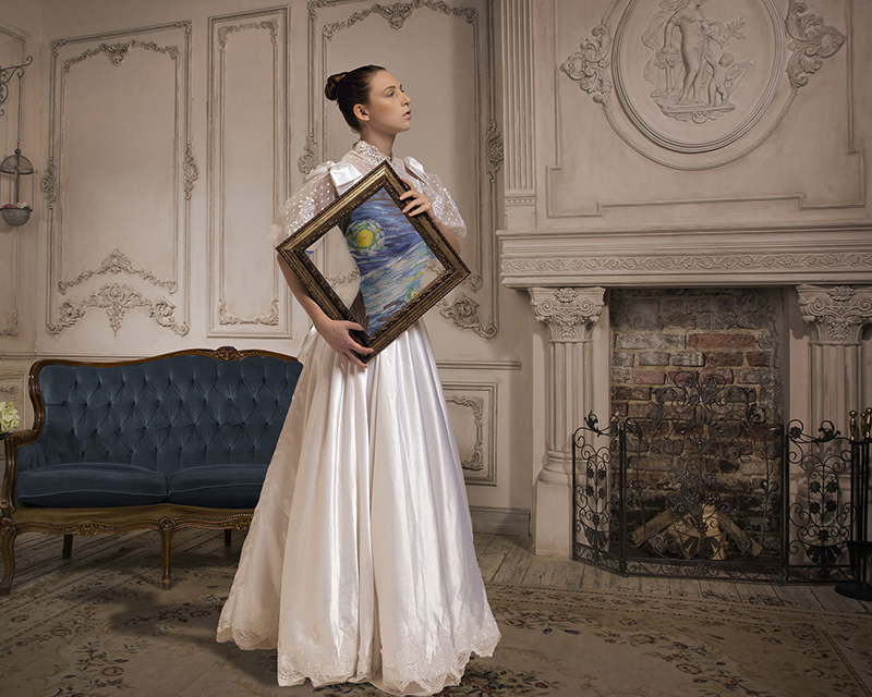 Woman in a white vintage dress holding a diamond-shaped frame displaying a Van Gogh style painting, standing in a room with ornate wall paneling, a blue velvet sofa, and a fireplace with decorative molding