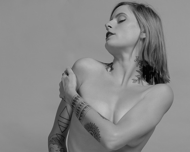 Woman with short hair and various tattoos on her neck and arm, posing with eyes closed and head slightly tilted back, one arm crossed over her chest.