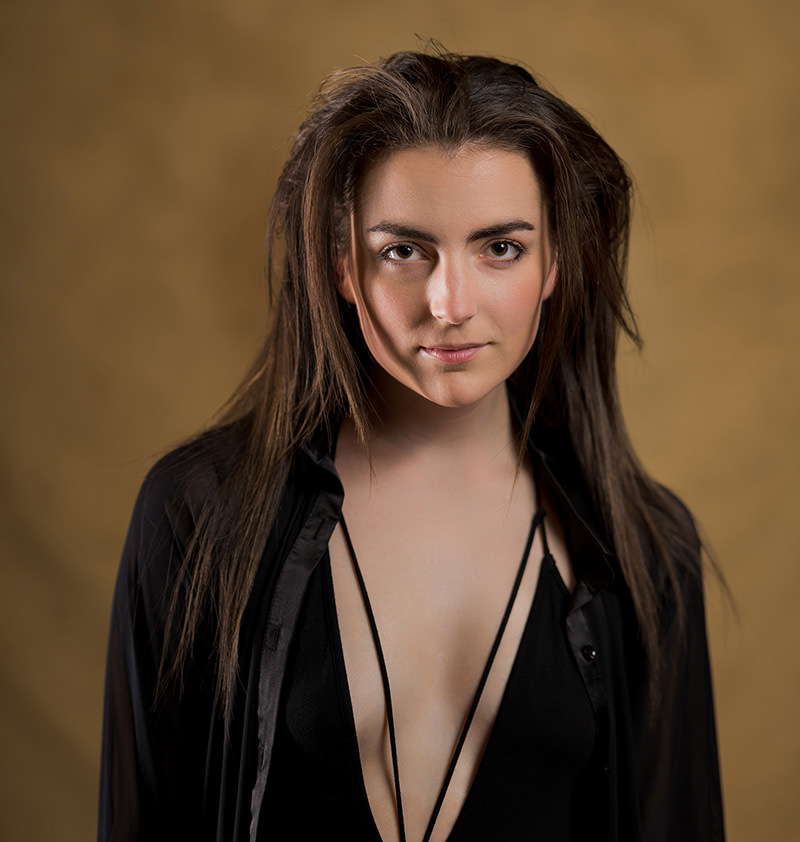 Young woman with straight brown hair wearing a deep v-neck black top and a black open shirt against a blurred brown background, looking directly at the camera with a confident expression.