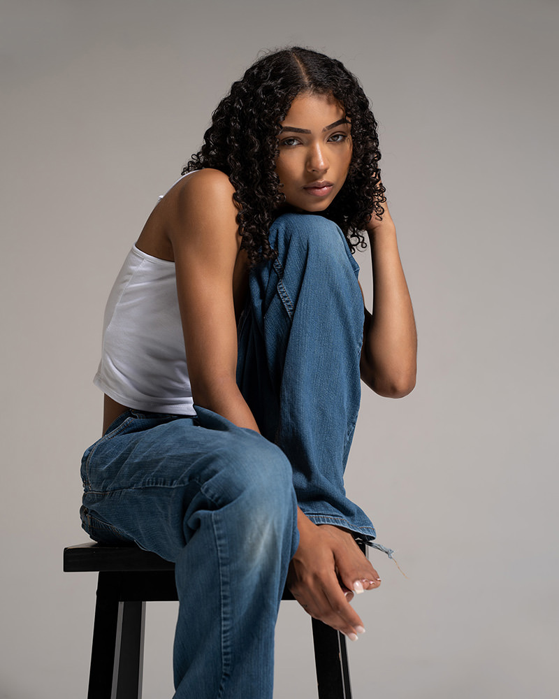 Young woman with curly hair sitting on a stool, hugging her knees close to her chest, wearing a white sleeveless top and blue jeans against a plain gray background