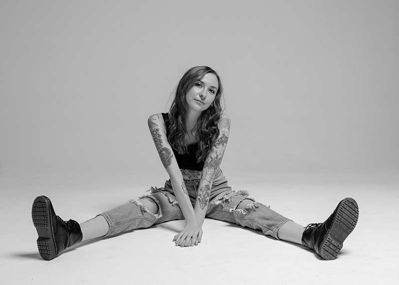 Young woman with long wavy hair, multiple tattoos on her arms, wearing a black sleeveless top, ripped jeans, and black boots, sitting on the floor with legs spread wide and hands touching in front, looking at the camera.