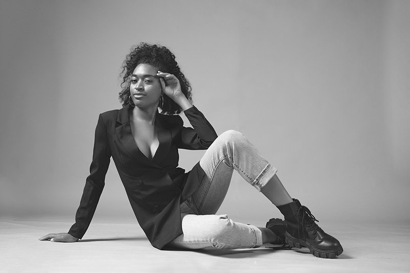 Woman with curly hair sitting on the floor, wearing a black blazer, light jeans, and black boots, looking confidently at the camera against a plain background
