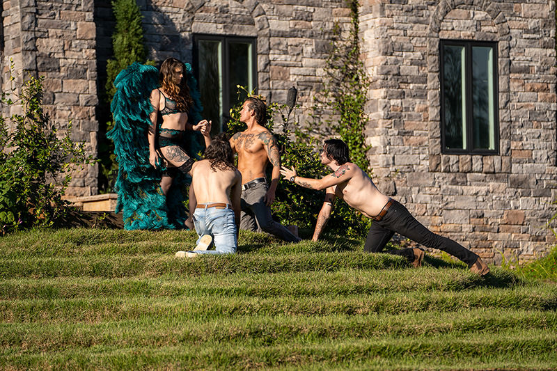 Three shirtless men with tattoos kneeling and reaching toward a woman standing on a grassy hill, dressed in black lace lingerie and large green feathered wings, with a stone building in the background.
