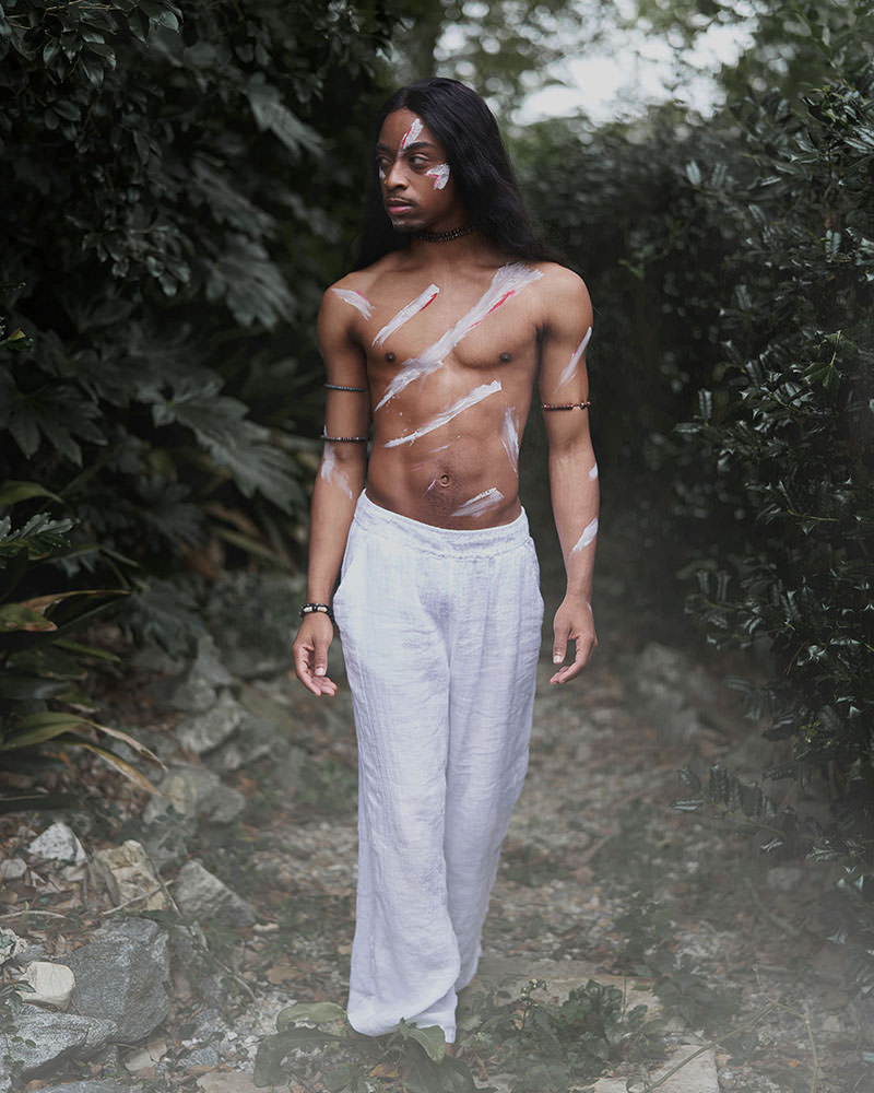 Bare-chested person with long hair and white and red body paint standing on a rocky, leafy path flanked by dense green foliage, wearing loose white pants and beaded bracelets
