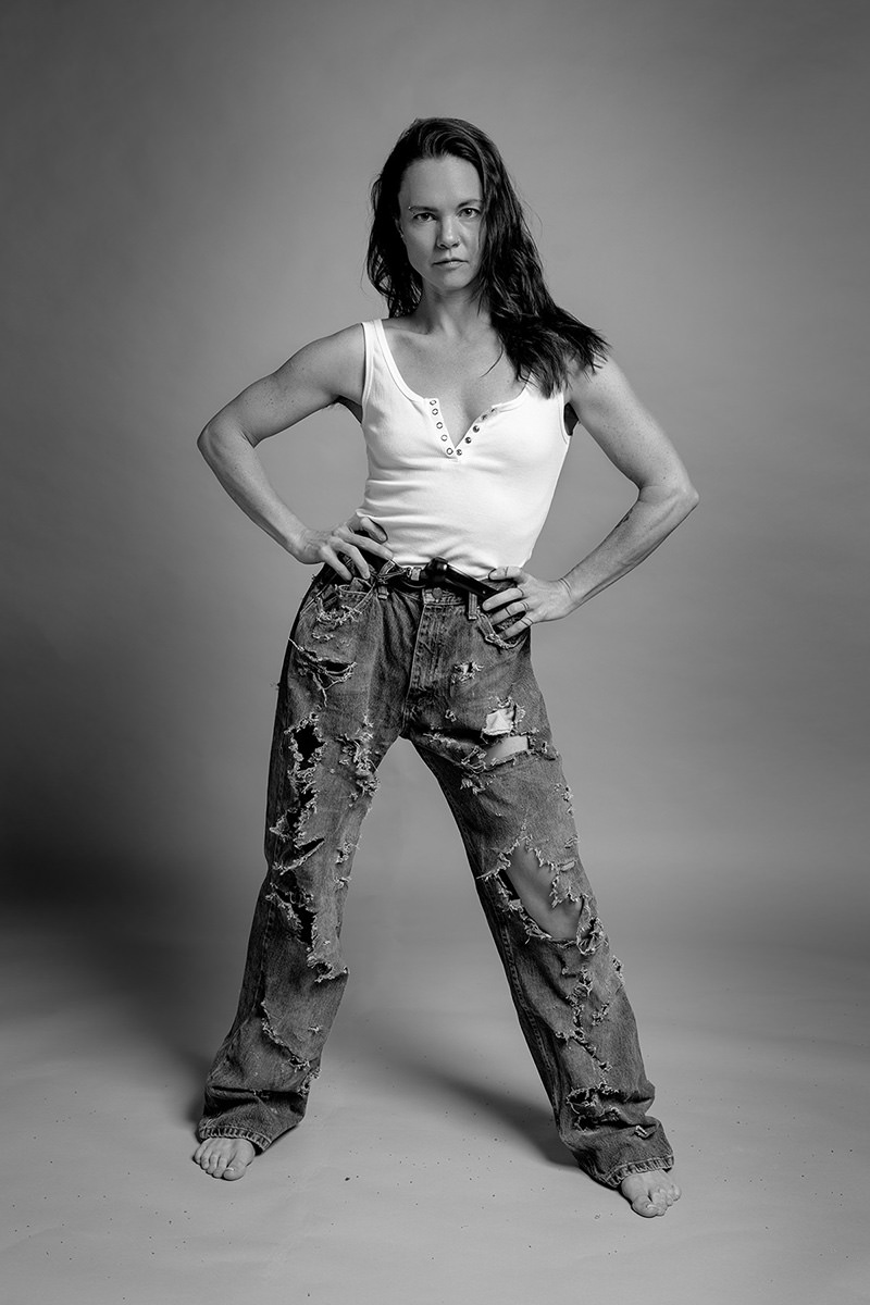Woman standing barefoot with hands on hips wearing a white sleeveless top and heavily ripped jeans against a plain backdrop