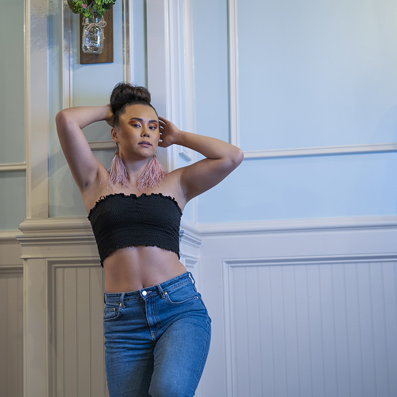Woman with dark hair styled in a bun, wearing a black strapless top, blue jeans, and large pink feather earrings, standing with hands behind her head against a light-colored paneled wall with a hanging flower arrangement.