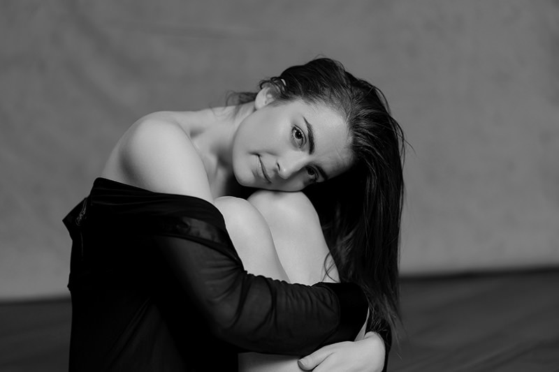 Young woman with long hair sitting curled up, resting her head on her knees, wearing an off-the-shoulder dark top, looking gently toward the camera against a blurred background