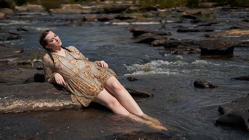 Young woman in a floral dress reclining on a rock with legs submerged in a shallow river, eyes closed, surrounded by flowing water and scattered rocks
