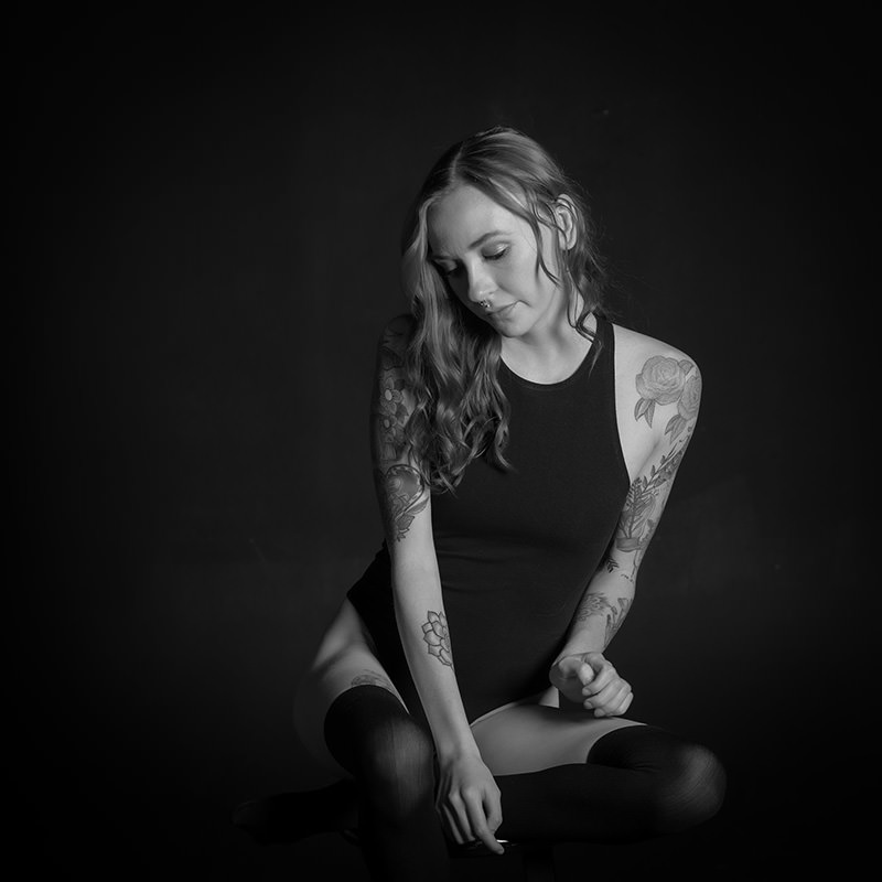 Young woman with long wavy hair wearing a sleeveless black bodysuit and thigh-high socks, sitting cross-legged against a dark background, with visible floral tattoos on both arms and a serene expression looking downward.
