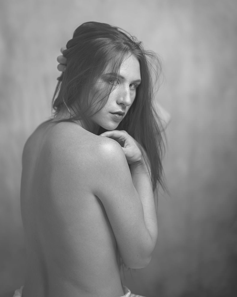 Young woman with long hair looking over her bare shoulder, hand touching her hair gently, against a softly blurred background in black and white.