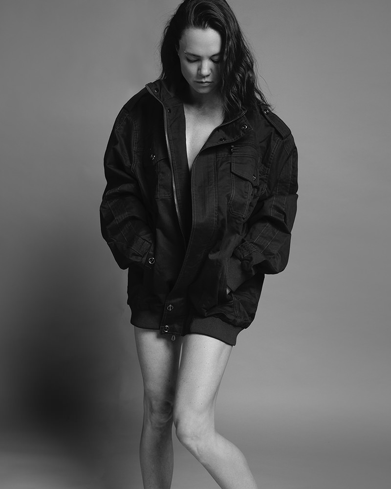 A person with long hair looking down, wearing an oversized dark jacket with hands in pockets, standing against a plain backdrop. The image is in black and white.