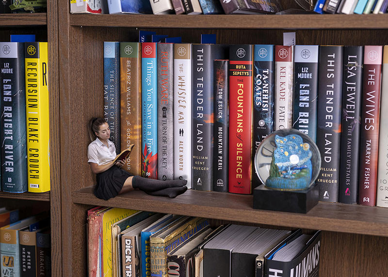 A girl dressed in a school uniform with long black socks sitting on a wooden bookshelf shelf, reading a book. She is surrounded by tall books standing upright, making her appear very small. There is also a snow globe nearby on the shelf.