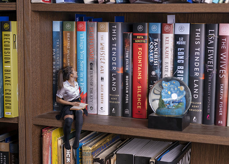 A small woman in a white blouse and black skirt sits on a bookshelf next to large books, reading a book herself. The bookshelf contains various colorful books and a small globe ornament.
