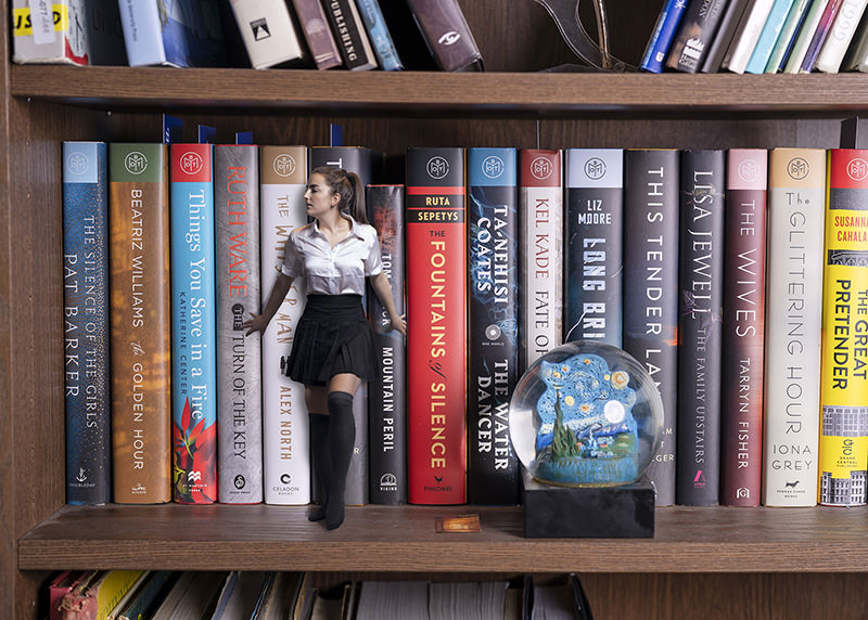 A tiny woman dressed in a white shirt and black skirt appears to walk between large upright books on a wooden bookshelf, blending size contrast with realism. To the right, a glass snow globe featuring a starry night scene sits on the shelf.