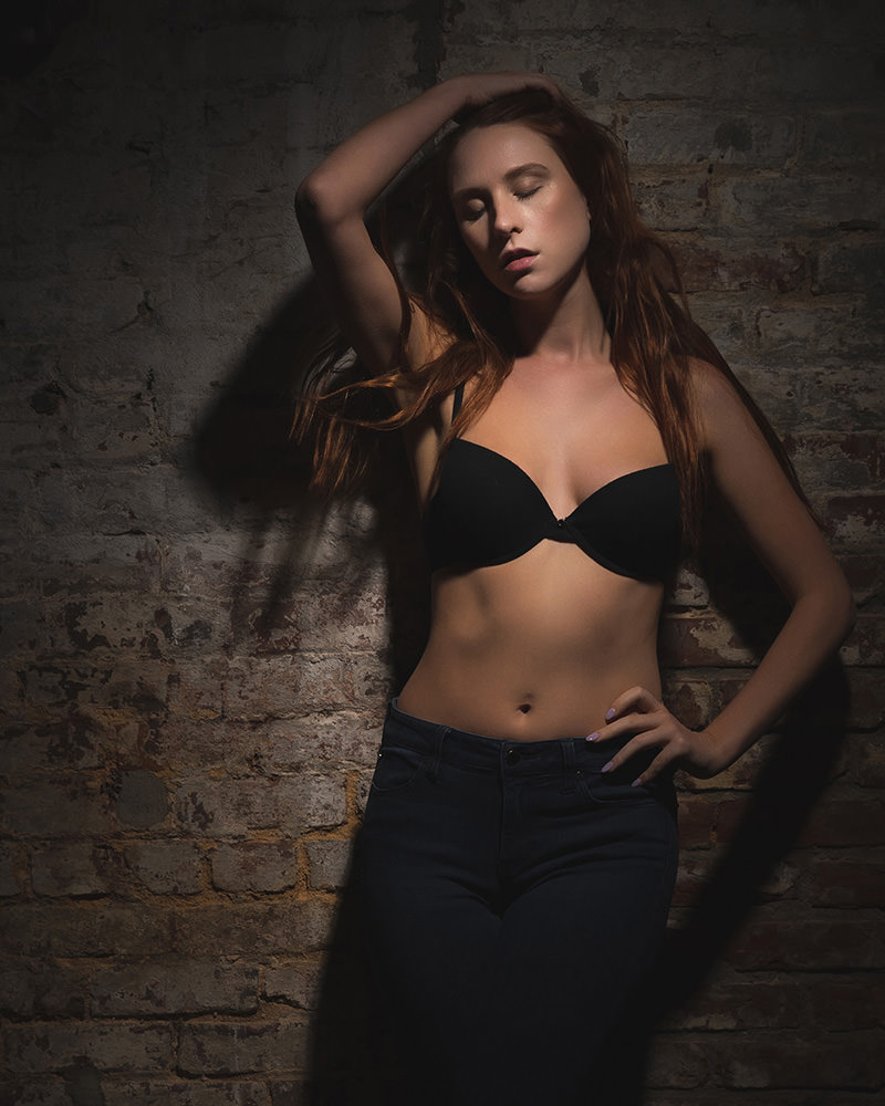 Young woman with long red hair wearing a black bra and dark jeans, posing with one hand behind her head and the other on her hip, standing against a textured brick wall with moody lighting highlighting her figure