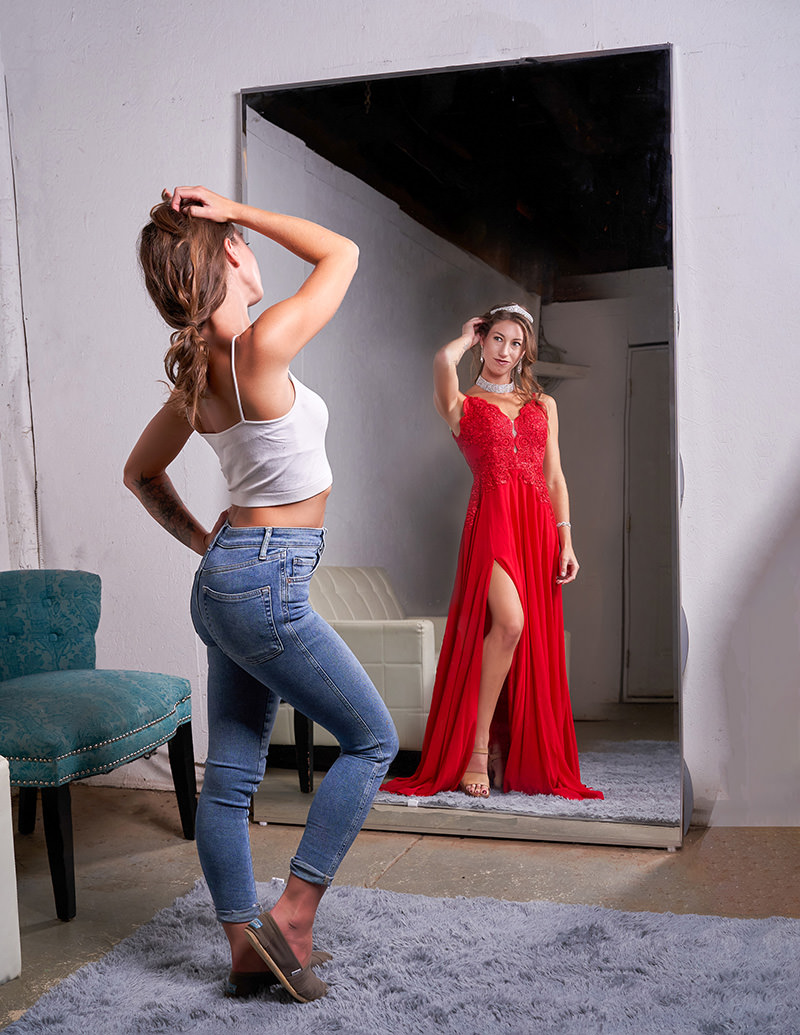 A woman in casual clothes stands in front of a large mirror, her reflection showing her wearing a glamorous red evening gown with a high slit, accessorized with jewelry and a headband.
