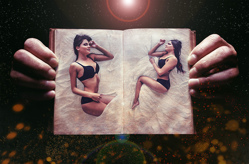 Two women in black lingerie lying on the pages of a large open book held by oversized hands, surrounded by a dark starry background with floating light particles.