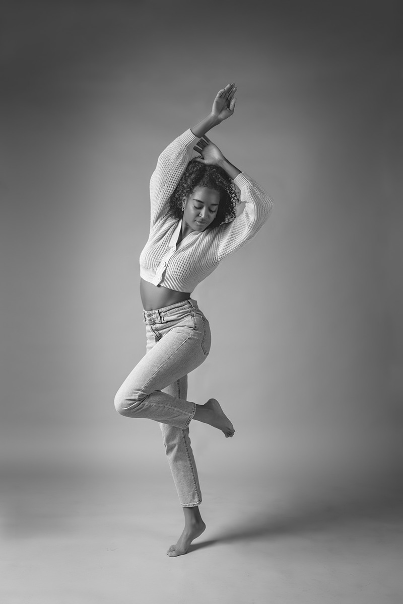 A woman with curly hair in a cropped sweater and jeans poses barefoot, balancing on one foot with arms raised and eyes closed against a plain backdrop