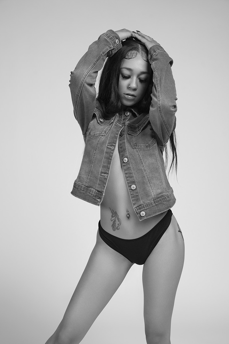 Young woman with long hair wearing a denim jacket and black underwear, standing with her arms raised above her head and eyes closed, featuring a unicorn tattoo on her side and a belly button piercing