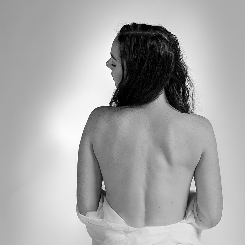 Back of a woman with long dark hair looking to the side, bare shoulders visible against a light gray background, wearing a white garment loosely draped around her lower back