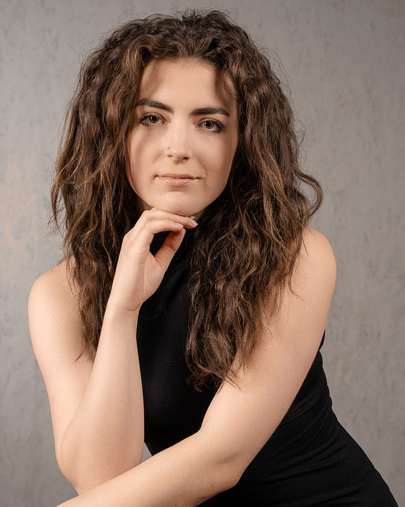 Young woman with long, curly brown hair wearing a sleeveless black dress, posing with her chin resting on her hand, against a plain gray background.