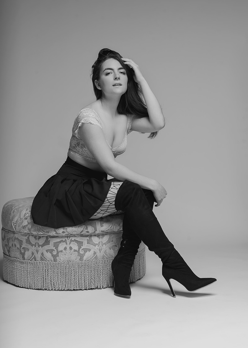 Woman with long dark hair seated on a patterned round ottoman, wearing a lace crop top, pleated skirt, fishnet stockings, and high-heeled knee-high black boots, with one hand touching her hair and a soft confident expression