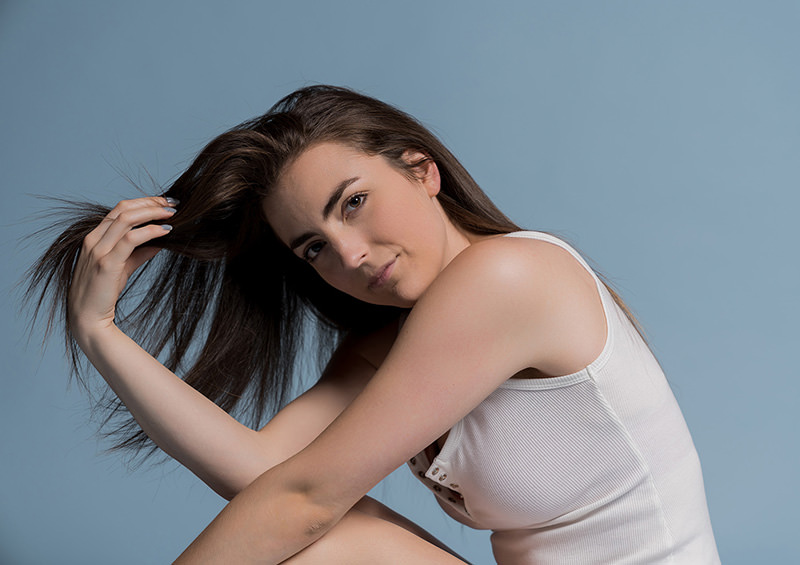 Woman with long brown hair wearing a white sleeveless top, sitting with her knees up and holding her hair with one hand against a solid light blue background