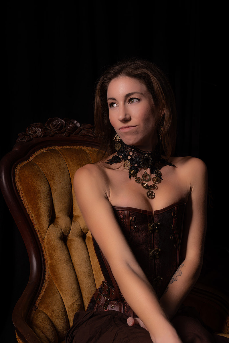 Woman in a brown leather corset and intricate black and gold steampunk necklace sitting on a vintage mustard yellow upholstered chair, looking contemplatively to the side against a black background