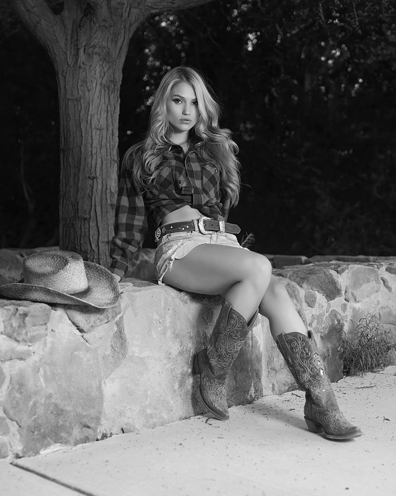 Blonde woman wearing a plaid shirt, denim shorts, and embroidered cowboy boots sitting on a stone wall with a cowboy hat nearby, set against a dark natural background.