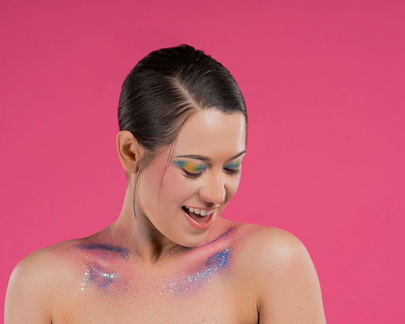 Young woman with slicked-back dark hair looking down with a joyful expression, colorful eyeshadow in blue, yellow, and green hues, and glittery, shimmering makeup on her shoulders against a bright pink background.